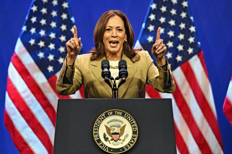 Vice President Kamala Harris speaks at a podium, with her index fingers on both hands pointed up.