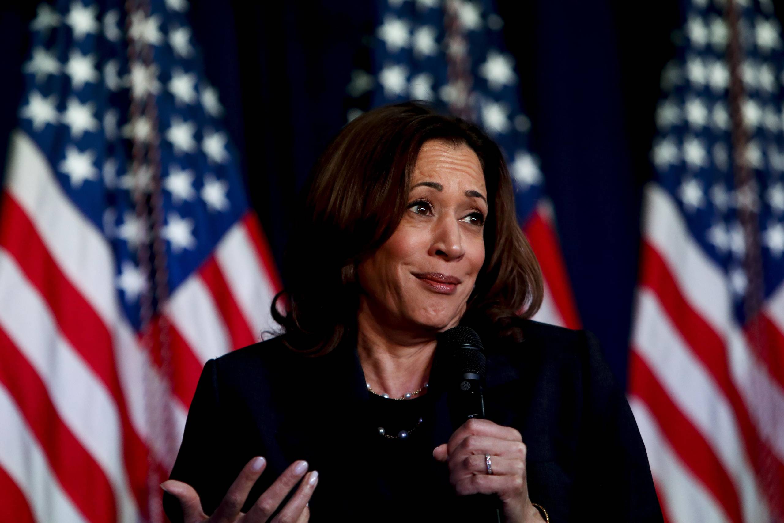 A woman wearing a black jacket holds a microphone with three U.S. flags in the background.