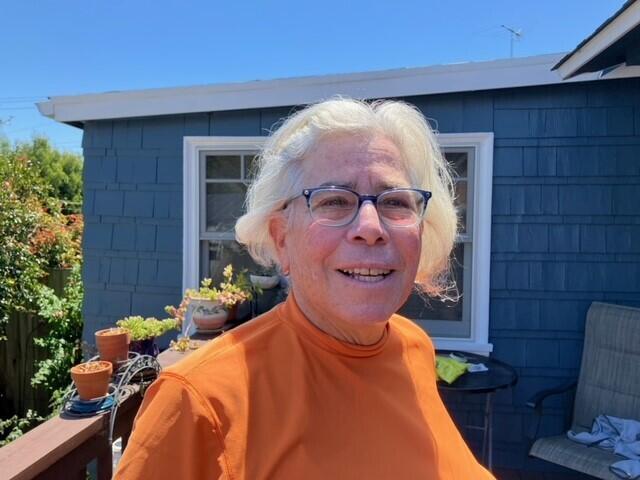 A woman with grey hair stands in front of a blue house wearing an orange sweater.