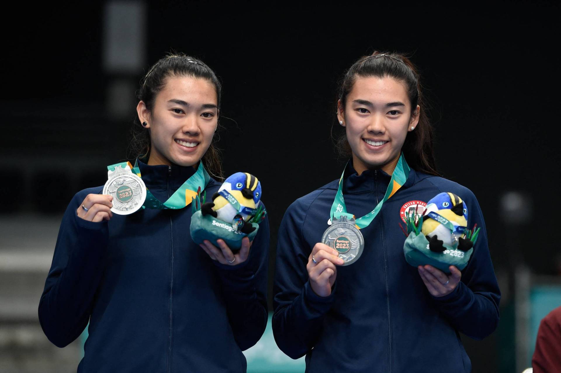 Two young women of Asian descent stand smiling together, holding up medals and commemorative toys.
