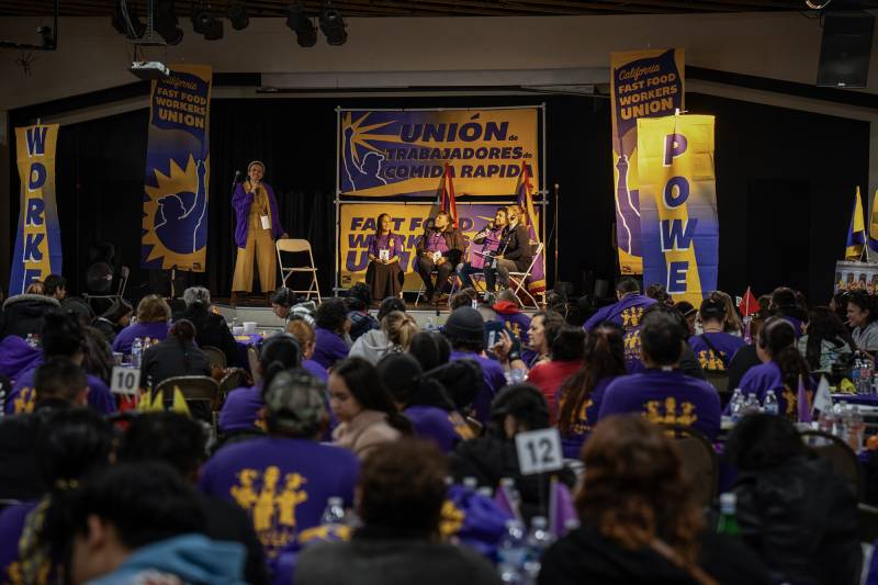 An audience watches a woman wearing purple and yellow holds a microphone with four other people seated near her on stage.