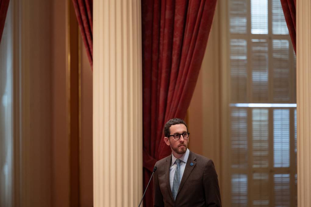 A white man wearing glasses and a business suit stands by a microphone inside a building.