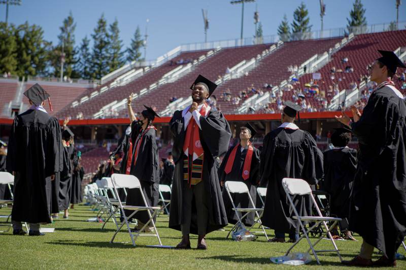 A young Black man wearing a black cap and gown interlocks his fingers, looking up as other young people wearing the same attire walk around. Everyone is in a stadium with white fold-up chairs on the field.