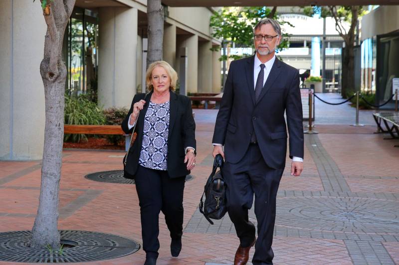 A middle-aged white woman with blond hair walks through a courtyard, next to a middle-aged man wearing a suit and carrying a briefcase.