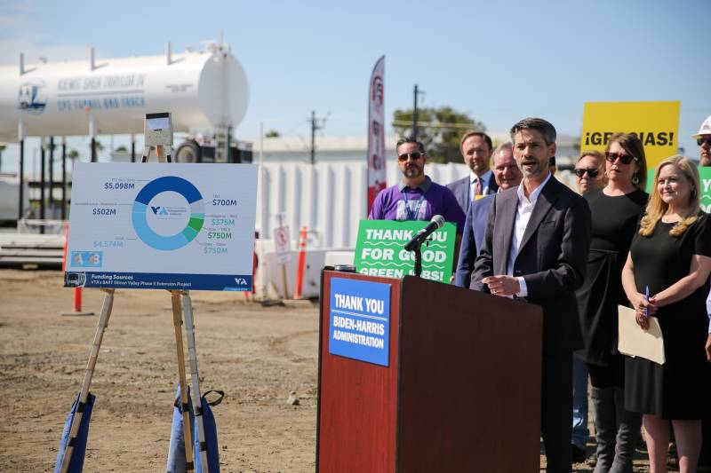 The mayor of San Jose stands behind a podium. A poster breaking down the project budget is displayed next to the speaker.