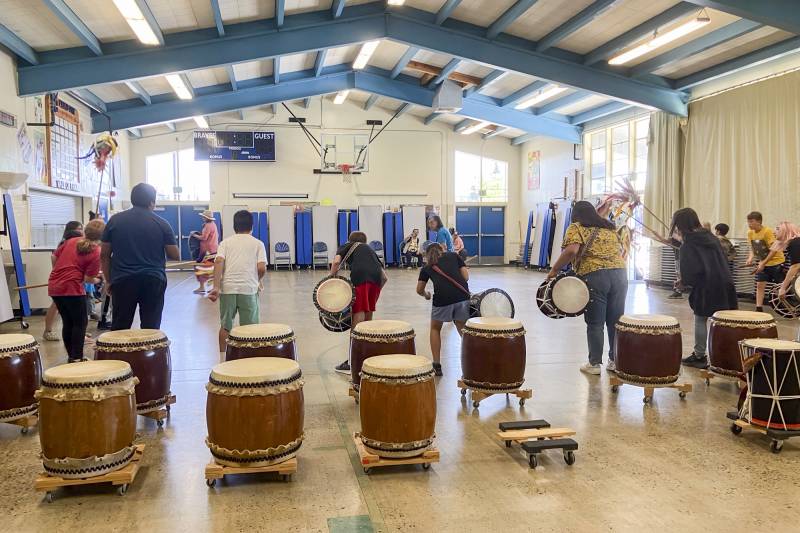 Several children and two adults hold and play large drums in a gym.