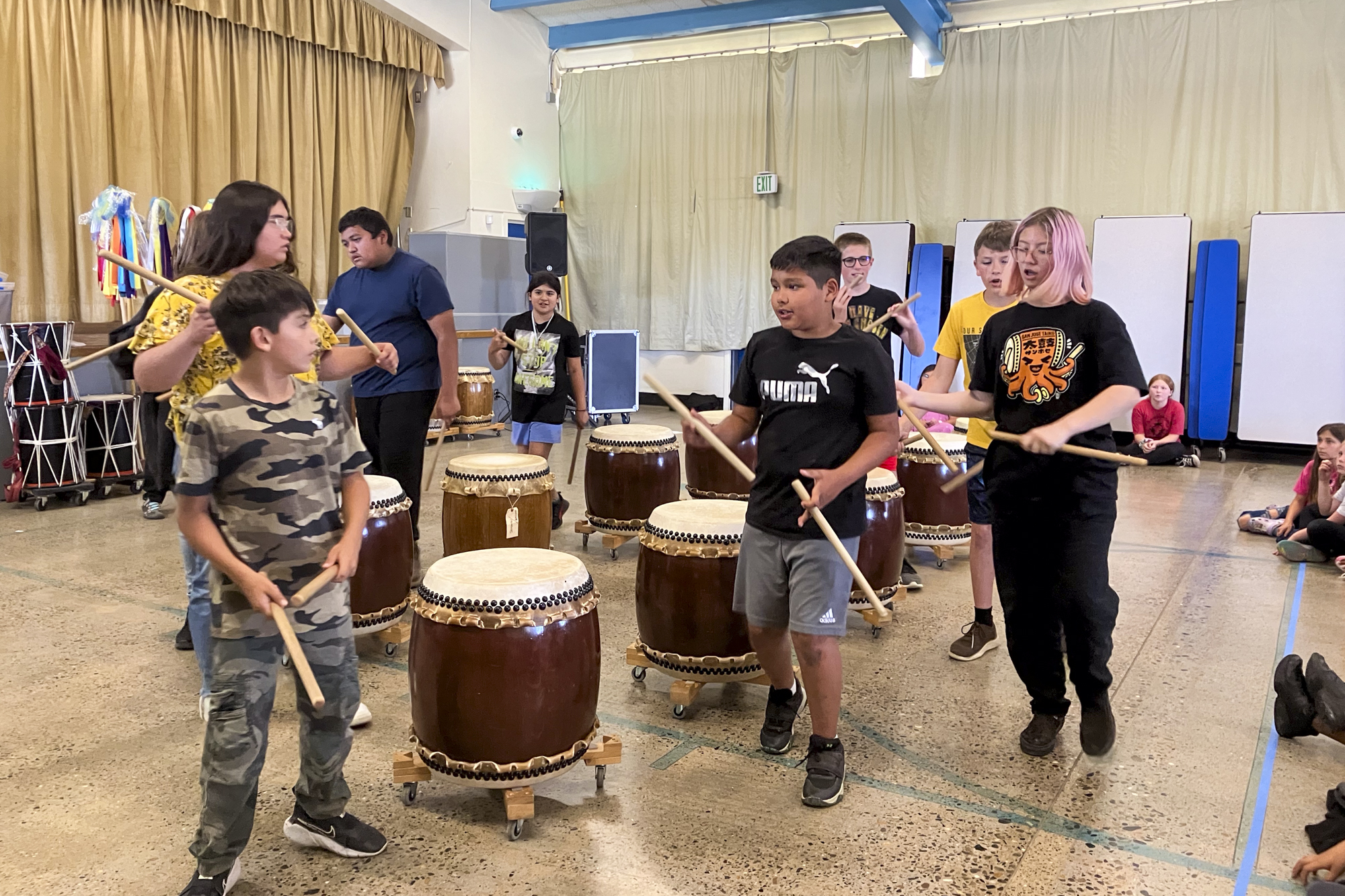 Children hold drumsticks near large drums in a gym.