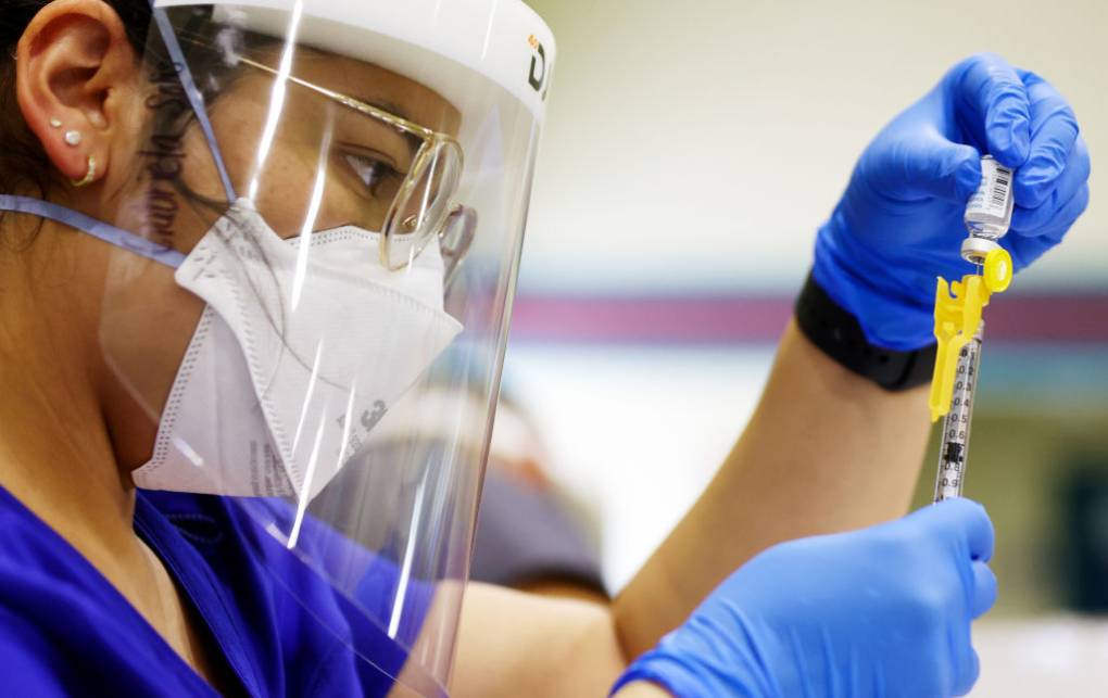 A woman wearing glasses, a face mask and a visor with blue surgical gloves holds a syringe and a small bottle.