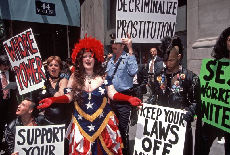 A buxom woman with voluminous curly hair in an American flag dress and elbow-length red opera gloves stands with a crowd of protesters. They're holding signs in support of decriminalizing sex work.