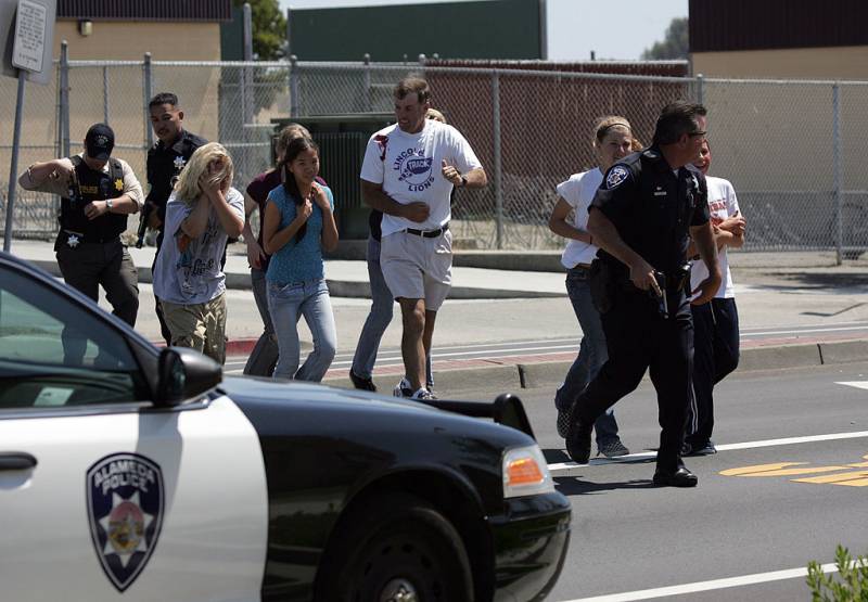 Three men dressed in police uniforms holding firearms walk with students and another man across the street.