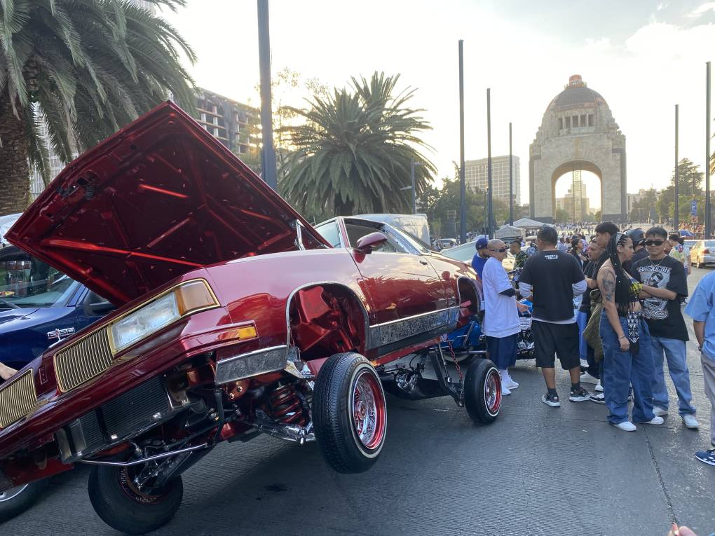 A small crowd on the street watches a red lowrider car that has one wheel in the air and the trunk open.