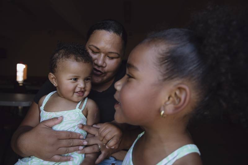 A woman holds a young child in her lap while another child looks in their direction.