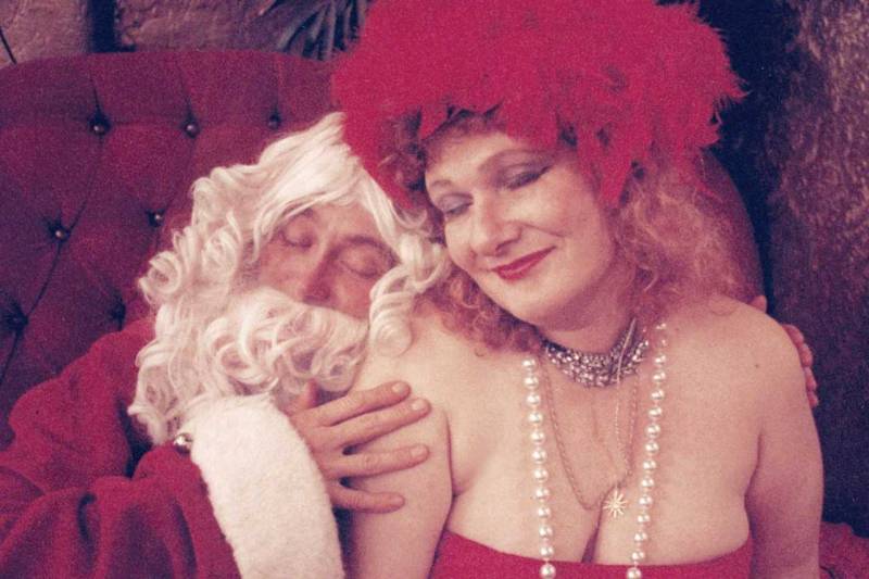 A woman in a strapless red dress with a red feathered hat cuddles up next to a man dressed as Santa Claus.