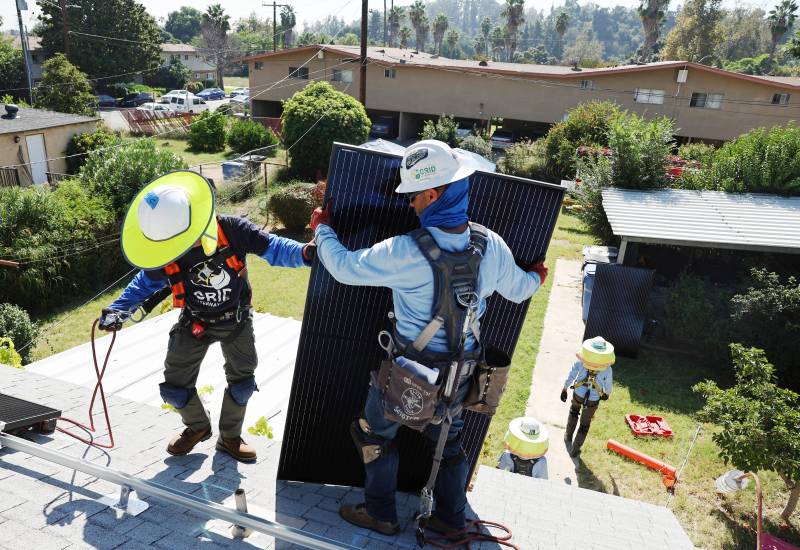 Two workers wearing hard hats carry a solar panel onto a rooftop.