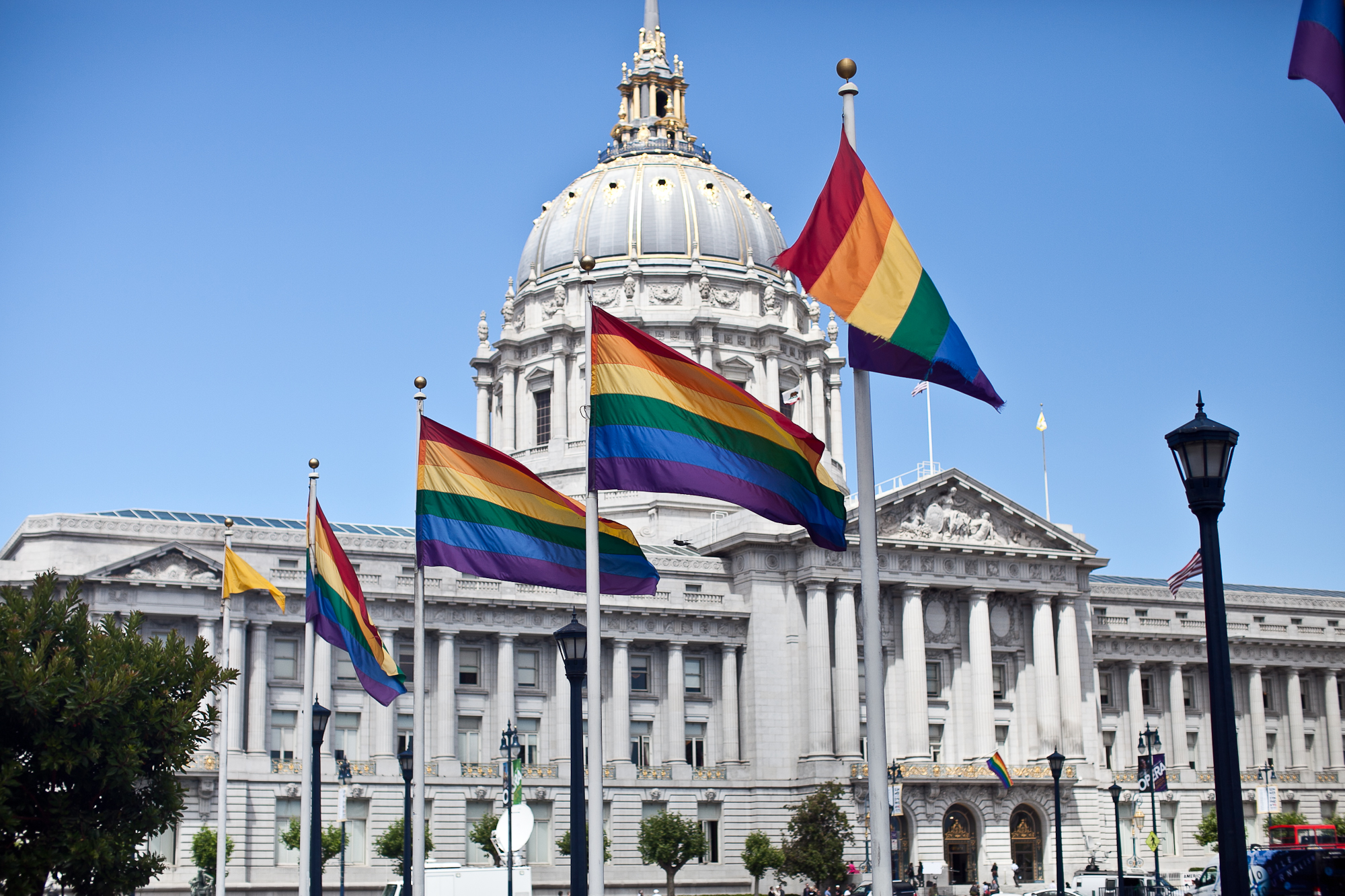 A row of flagpoles displaying rainbow flags stands in front of San Francisco City Hall.