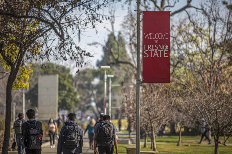 Young people wearing backpacks walk on a campus near a red sign that reads "Welcome to Fresno State."