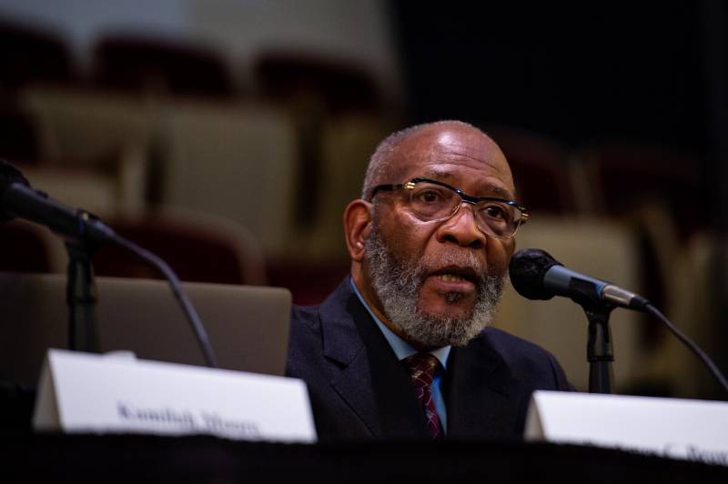 A Black man wearing glasses and business suit sits in front of a microphone inside a building.