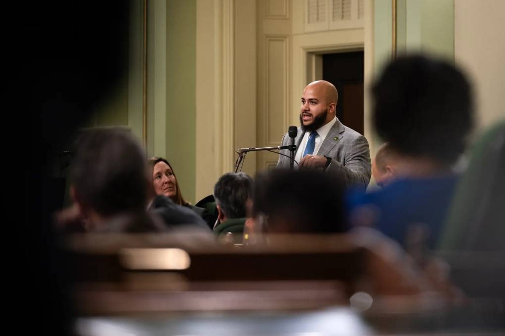 A young, bald Black man with a beard, wearing a grey suit, speaks at a formal hearing.