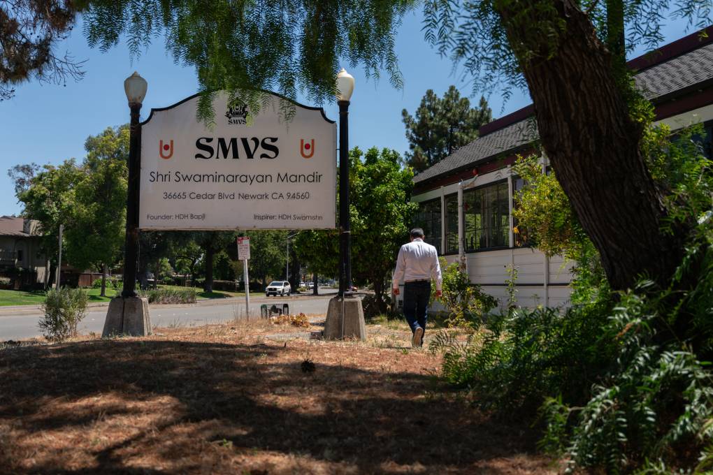 A man wearing a white shirt walks away from a sign that reads "SMVS Shri Swaminarayan Mandir" next to a building.