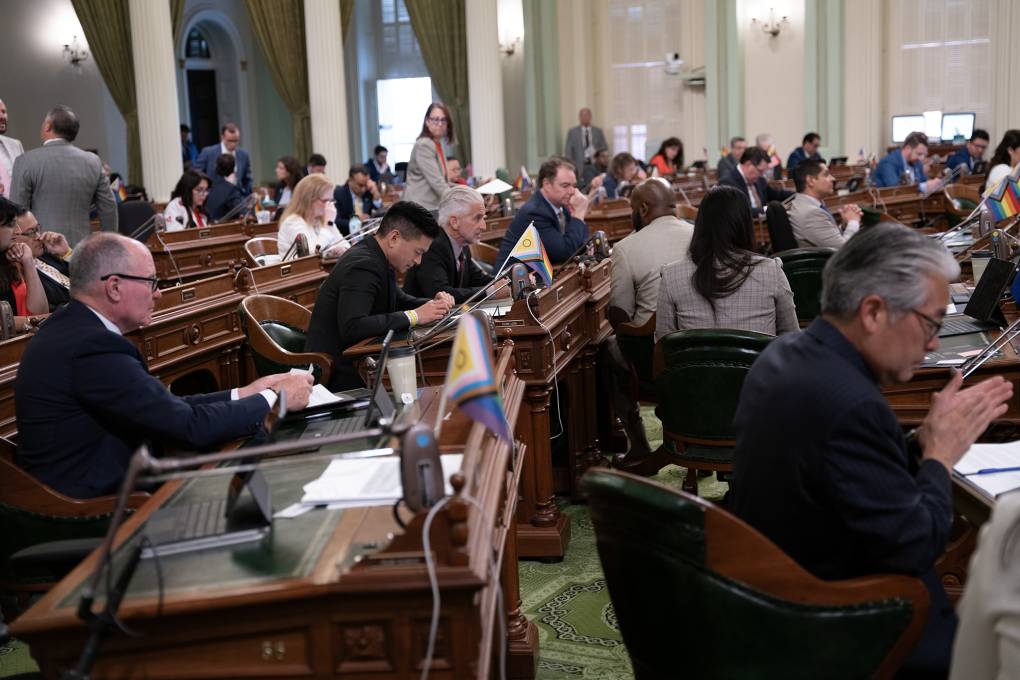 Several people seated in an assembly room with chairs, desks, small flags and microphones.