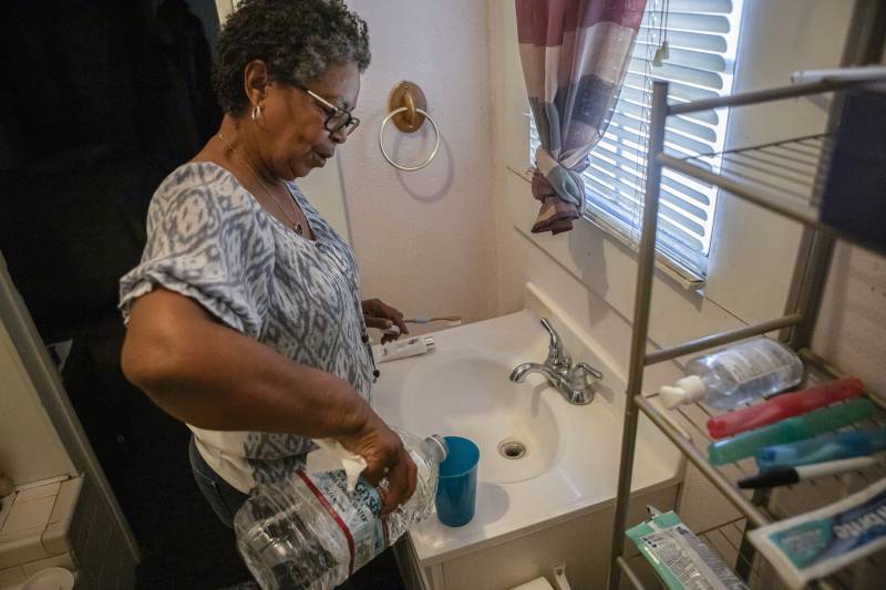 A Black woman wearing glasses and a patterned gray shirt pours water into a cup on a bathroom sink.