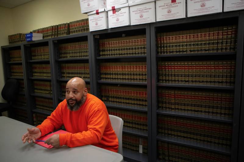 A Black, bearded, incarcerated man in an orange shirt sits at a table in front of a bookcase of legal books.