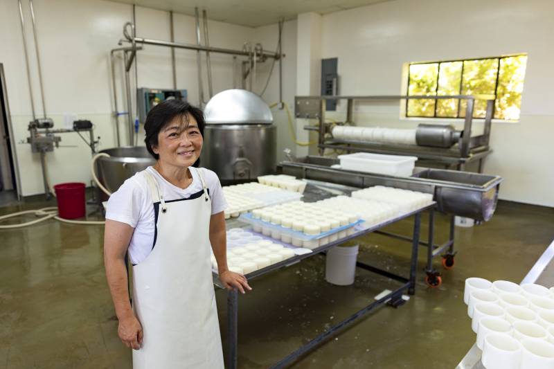 An Asian woman in a white apron stands in an industrial kitchen.