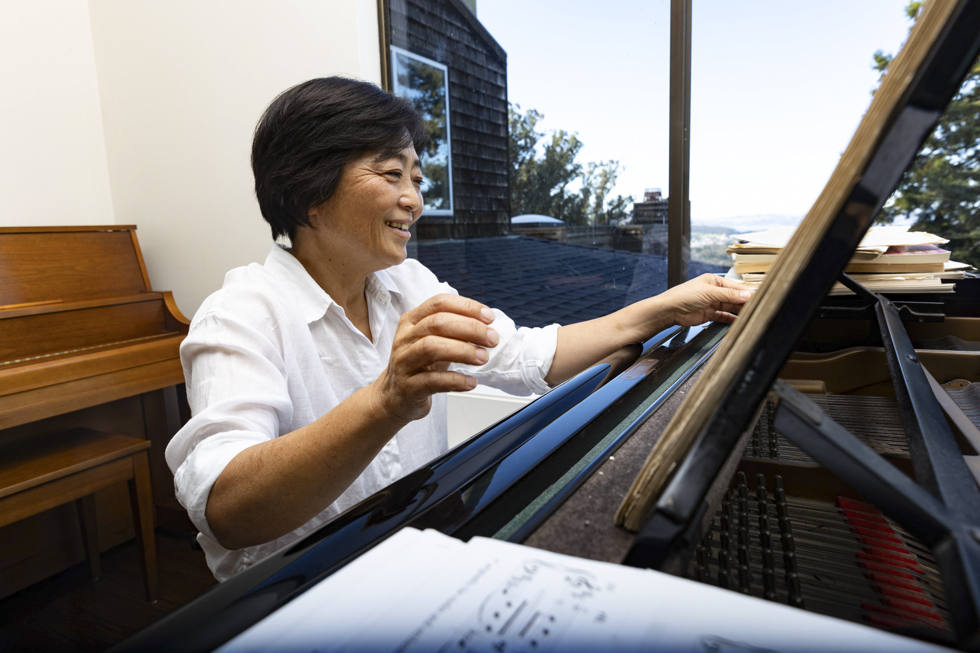 An Asian woman sits at a grand piano next to a window.