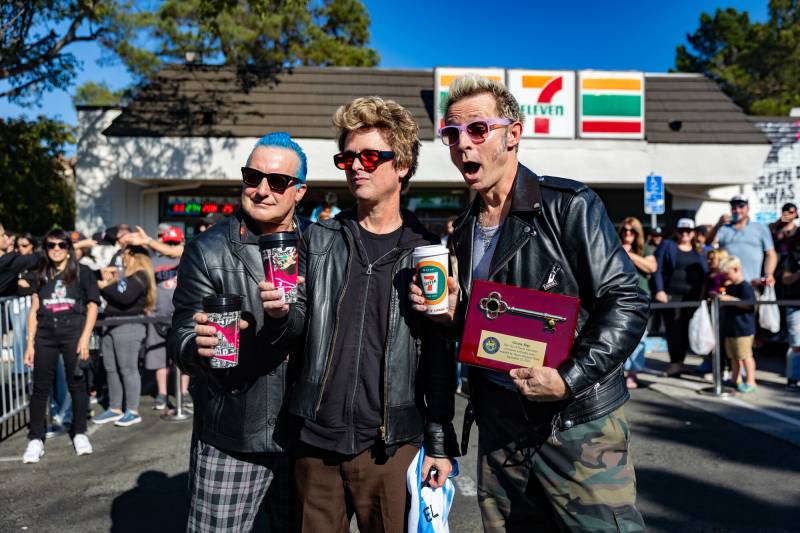 Three men wearing black jackets and sunglasses hold coffee cups and a plaque with a key on it in front of a 7-Eleven store.