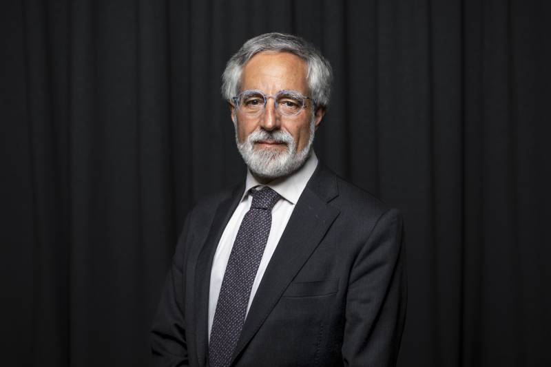 A white man with gray hair and a gray beard, wearing glasses and a navy suit, poses for a photo.