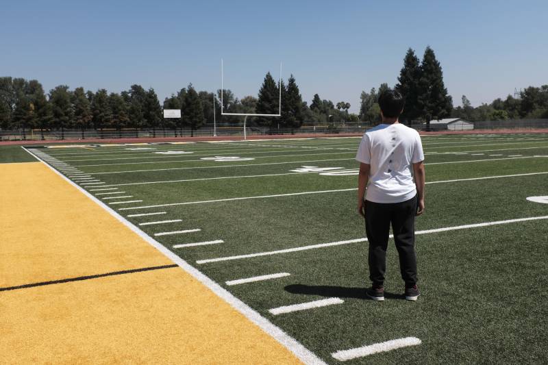 A young man wearing a white shirt and black pants stands on a football field.