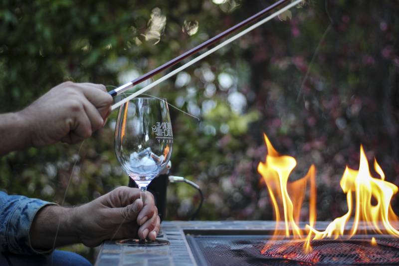 A hand holds a wine glass and a violin bow near an open flame outside.