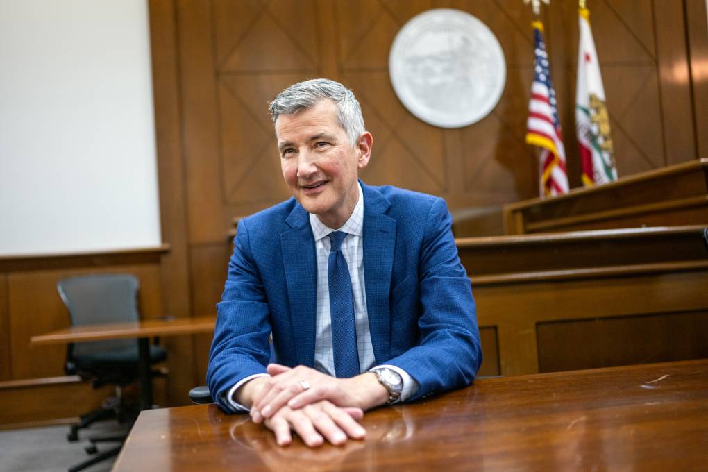 A white man wearing a blue dress suit sits at a table inside a courtroom.