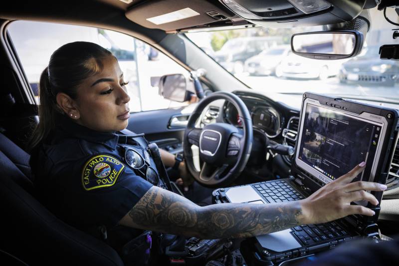 A woman dressed in a police uniform sits in a police vehicle and touches the screen of a laptop.