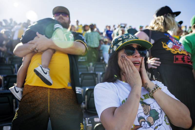 A woman with an A's hat has her hands on her cheeks, as a man behind her holds a small child, while standing amid a large crowd at a baseball stadium..