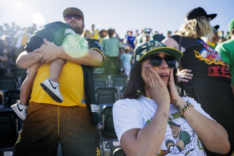 A woman with an A's hat has her hands on her cheeks, as a man behind her holds a small child, while standing amid a large crowd at a baseball stadium.