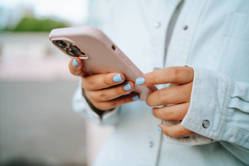 Close-up female hands with a blue manicure using pink smartphone outdoors.