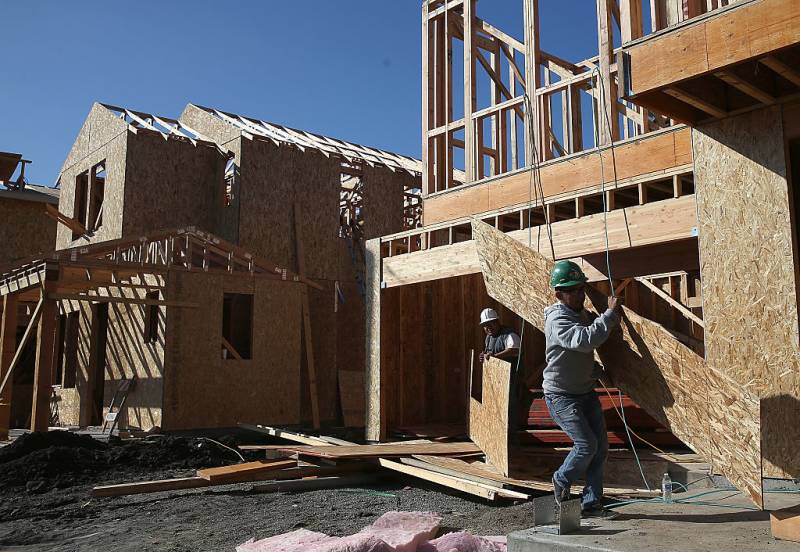 Two men dressed in construction uniforms carry large pieces of wood at a construction site for a house.