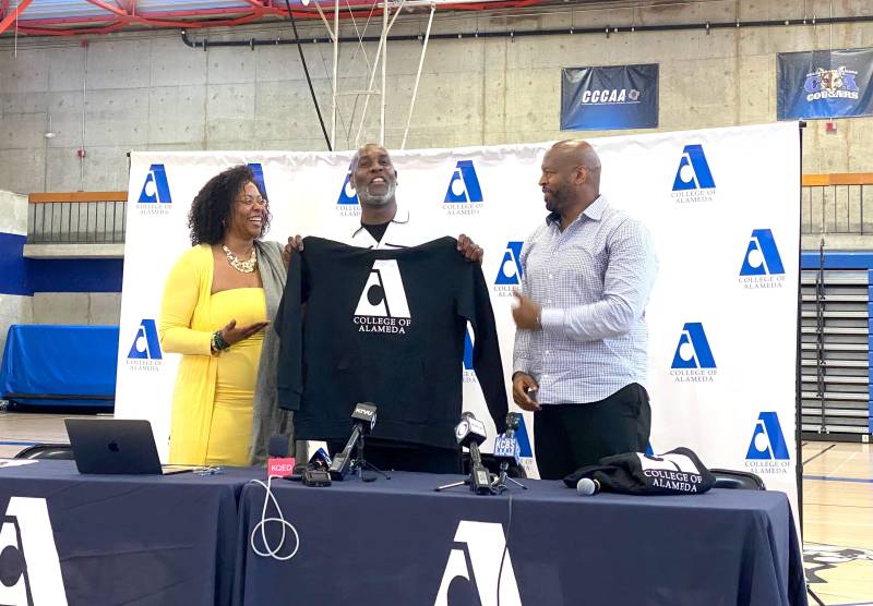 A Black man holds up a College of Alameda shirt, while standing between a woman and another man at a press conference.