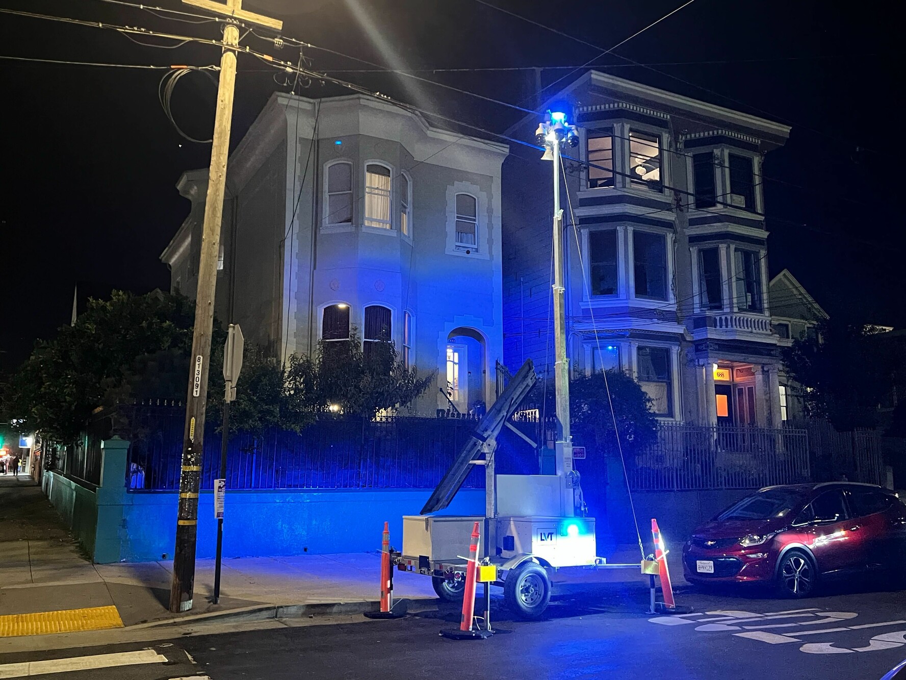 A 22-foot-tall contraption with three cameras and blue strobe lights sits next to a telephone pole at night. A blue light makes the houses in the background glow.