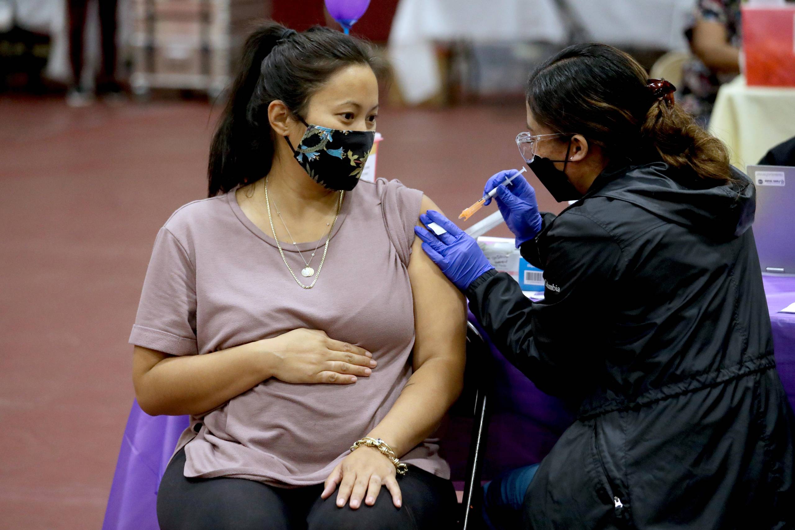A woman wearing a face mask sits next to another woman wearing a face mask and gloves while pushing a syringe needle into her arm.