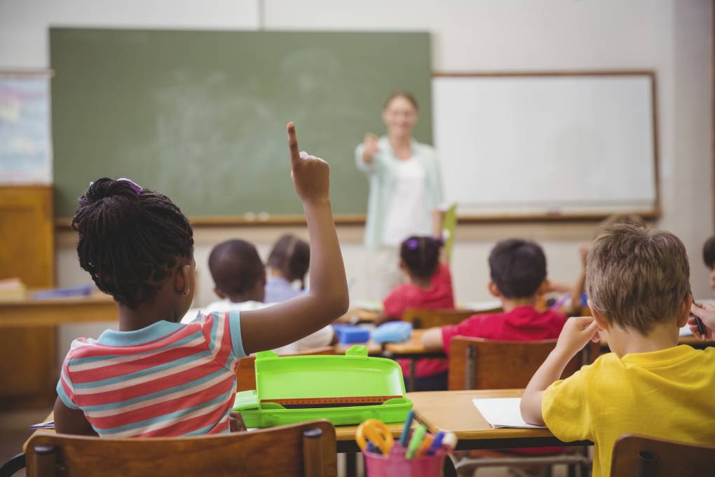 A view from the back of a classroom full of children. One little girl raises her hand as a teacher standing at the head of the class points towards her.