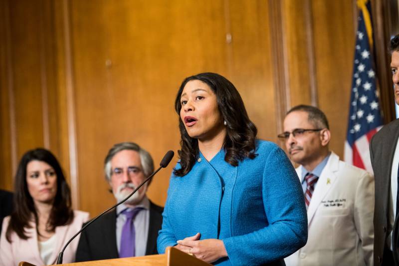 A Black woman wearing a blue suit stands in front of a podium with a microphone with three men and a woman behind her.