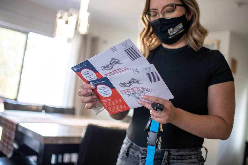 A woman wearing glasses and a black T-shirt with a facemask on looks down at two ballot envelopes.