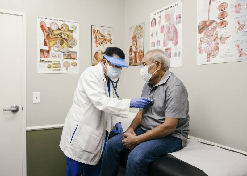 A man wearing a doctor's uniform holds an instrument to another man's chest who is sitting on a table.