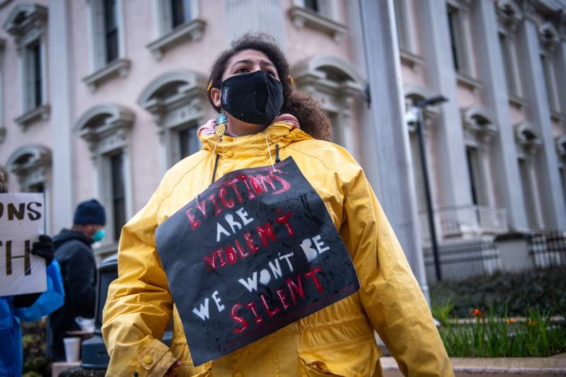 A woman wearing a yellow coat with a sign around her neck that reads "Evictions Are Violent. We Won't Be Silenced."