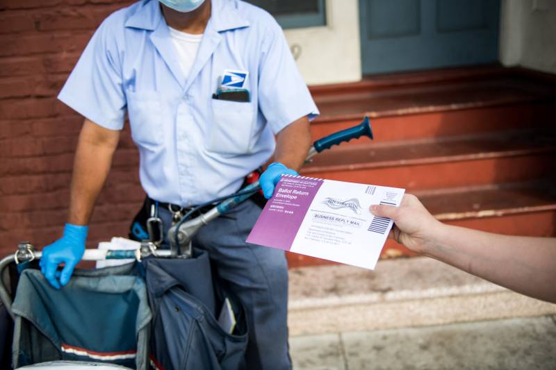 A postal worker on the street, wheeling a wagon, is handed a mail-in ballot.
