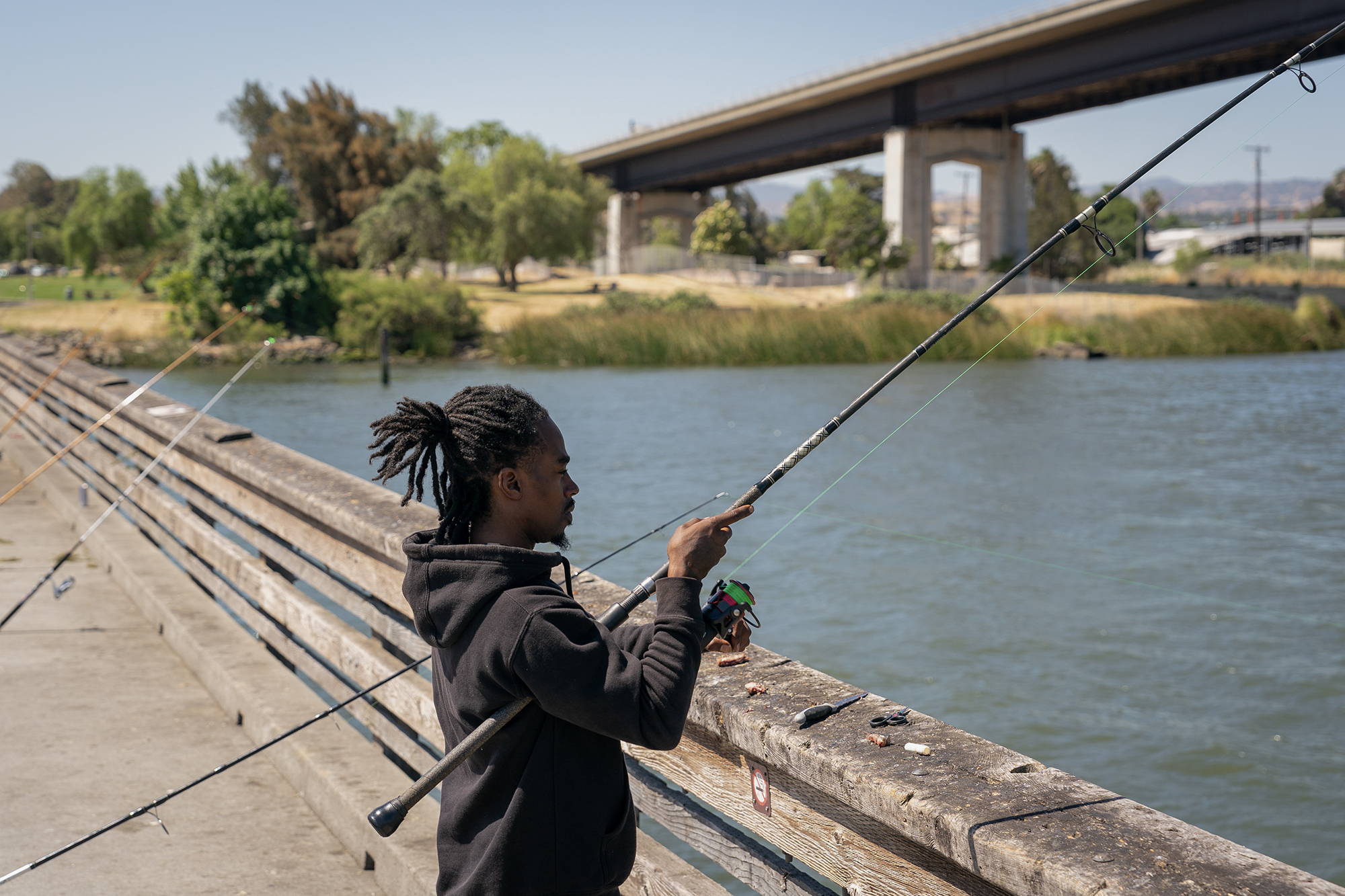 A Black man wearing a black hoodie holds a fishing rod over the side of a pier and water.