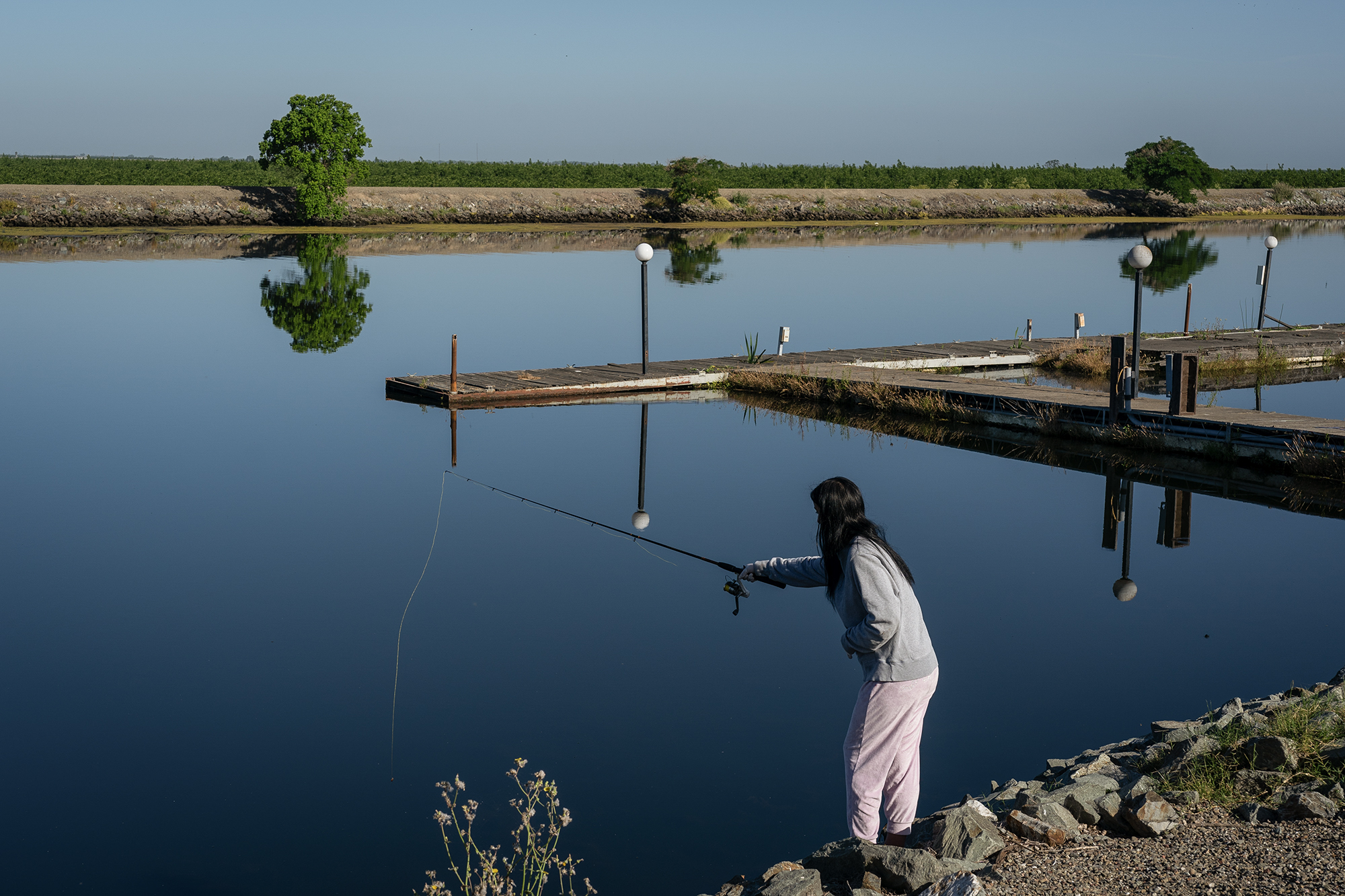 A woman wearing a gray shirt casts a fishing line out from the pole she is holding into a body of water.