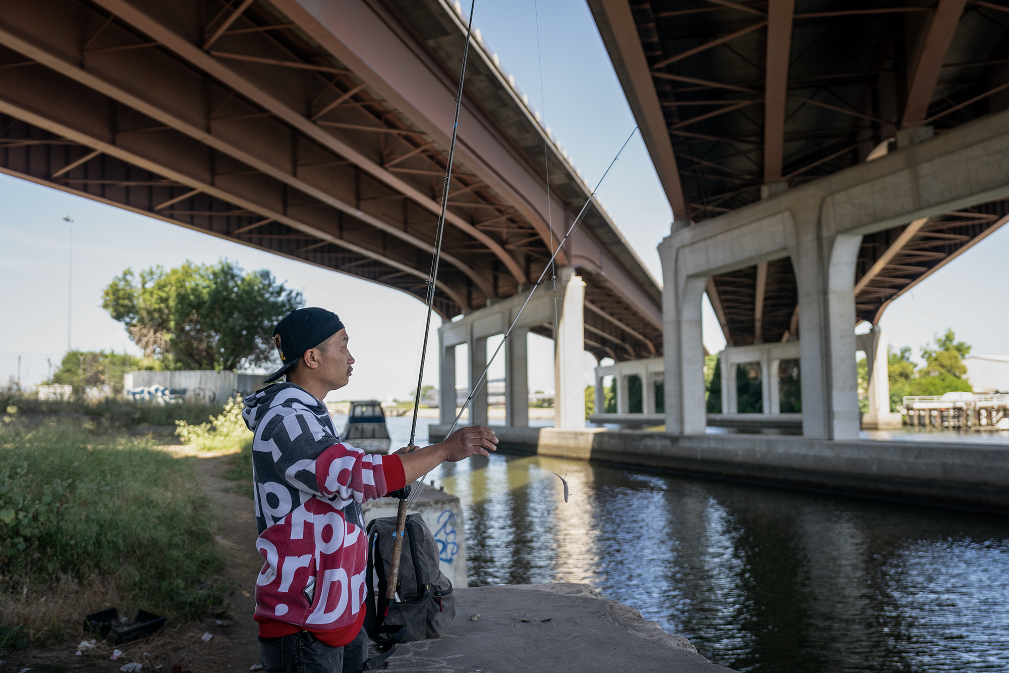 A man wearing a cap backwards and a grey and red shirt with white designs hold to fishing rods underneath an overpass.